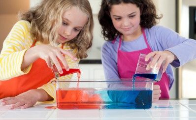 Two young people are pouring red and blue liquids into a glass pan.