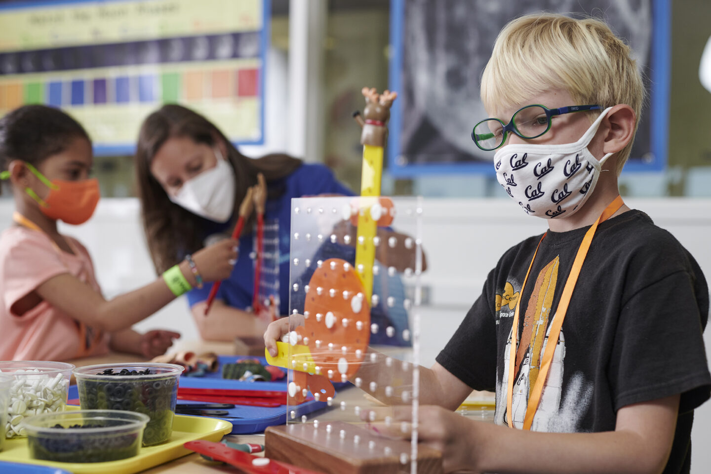 A child is constructing a project during the Toy Builders summer camp session and a teacher and student work on a separate project in the background.