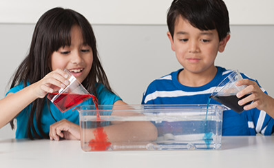 Two children pouring liquids into a glass pan.