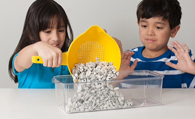 Two children pouring small stones into a glass pan.