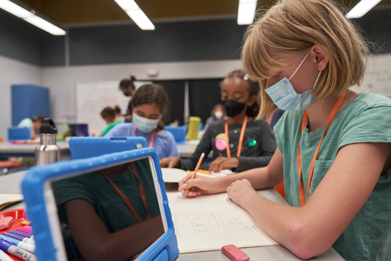 A camper is writing observations from a science activity and two more campers are writing in the background