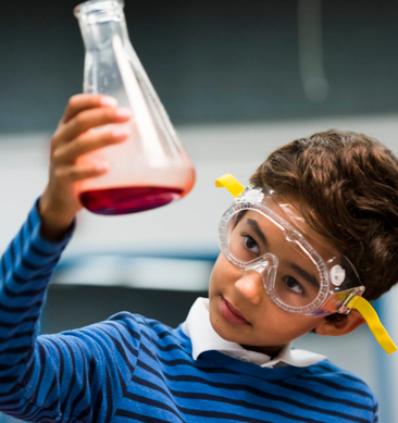 A young person wearing safety googles is holding up a science beaker that contains red liquid