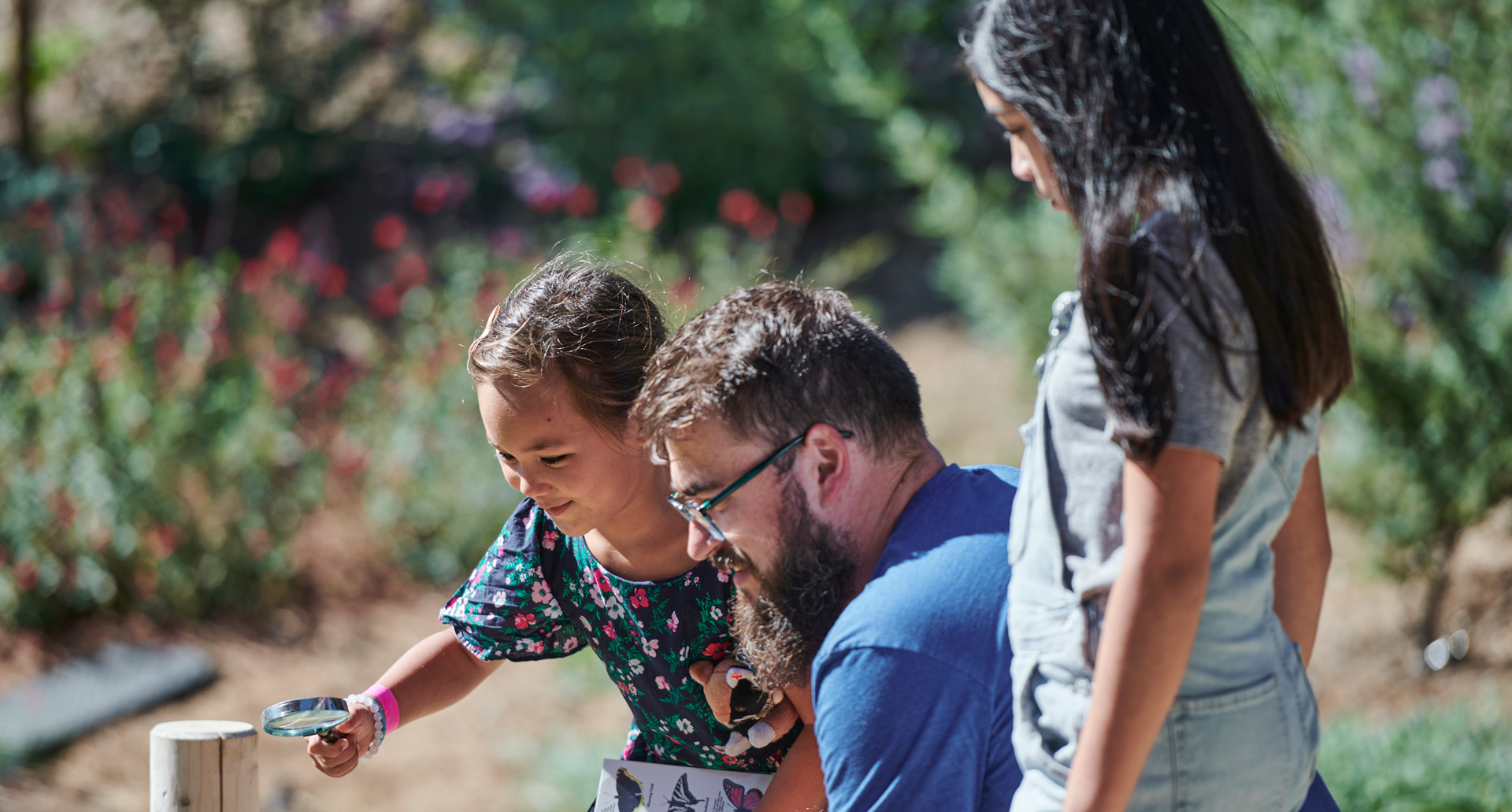 Two children and an adult are examining nature in the Outdoor Nature Lab using a hand lens.