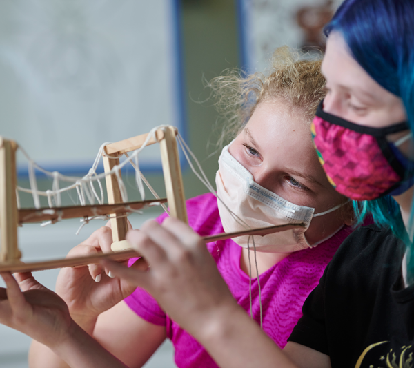 A student and an adult are holding up a model bridge that they have built.